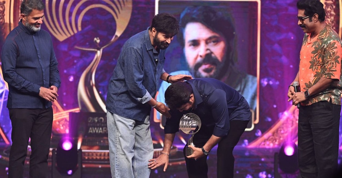 Mohanlal bends down to touch Mammootty’s feet at the Mazhavil Entertainment Awards 2026. Jayant Mammen Mathew, Executive Editor and Director of Malayala Manorama, also joined them on stage to present the awards. Photo: Jibin J Chempola/ Manorama