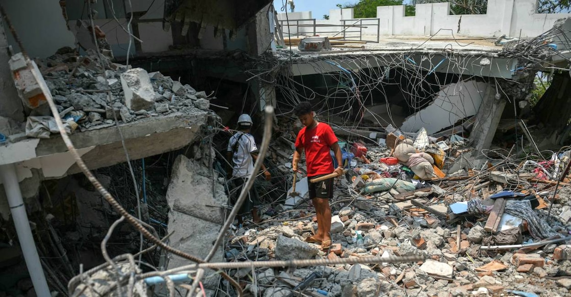People stand next to a destroyed building as rescuers look through the rubble to find survivors in Mandalay. Photo: AFP