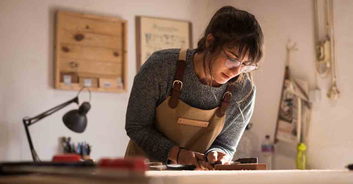 stay-at-home-2 A young female carpenter working as wood designer in a small carpentry workshop. Young business woman handcrafting a piece of timber and designing new house furniture. Entrepreneurs concept lifestyle